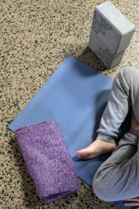 Photo of a person sitting on a blue yoga mat next to a purple towel and grey yoga blocks