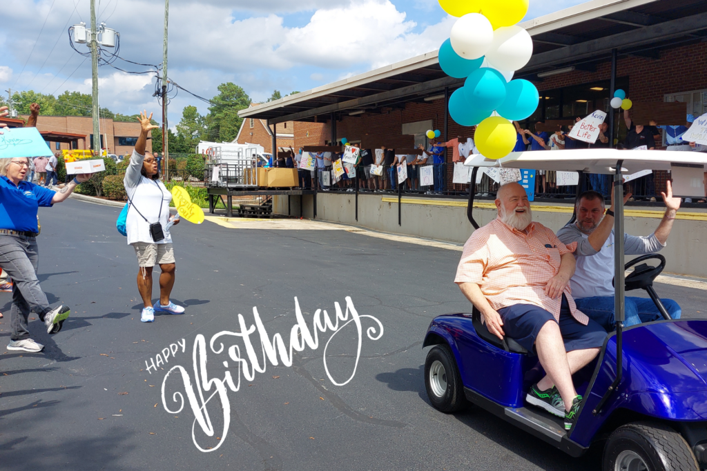 Kevin Mcdonald riding in a golf cart with TROSA residents cheering him on and displaying handmade signs that thank him and celebrate recovery