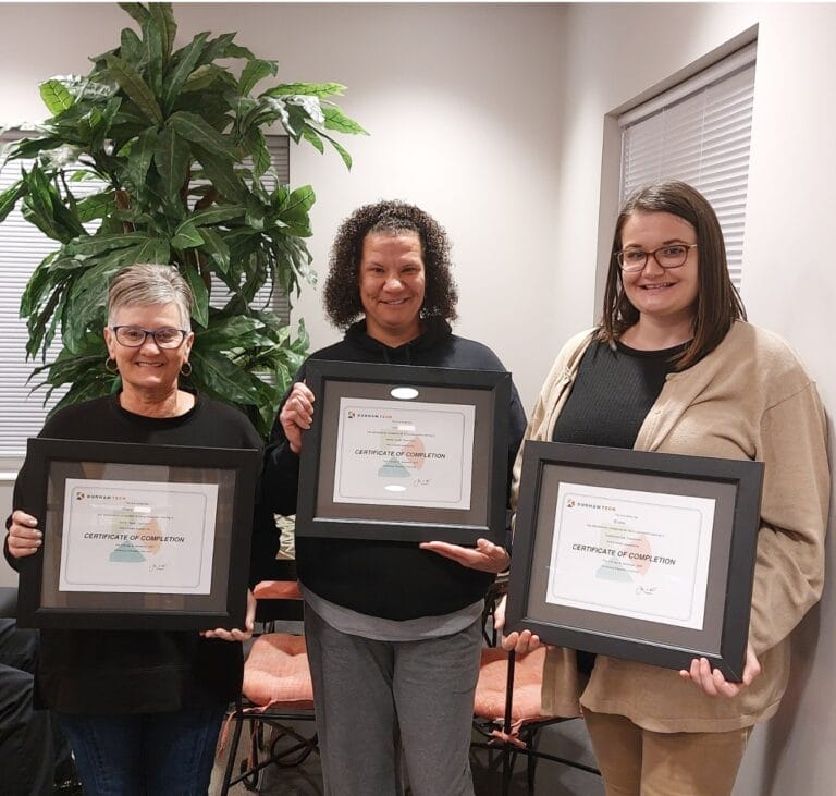 three female TROSA residents stand next to each other, smiling, and hold Durham Technical Community College certificates