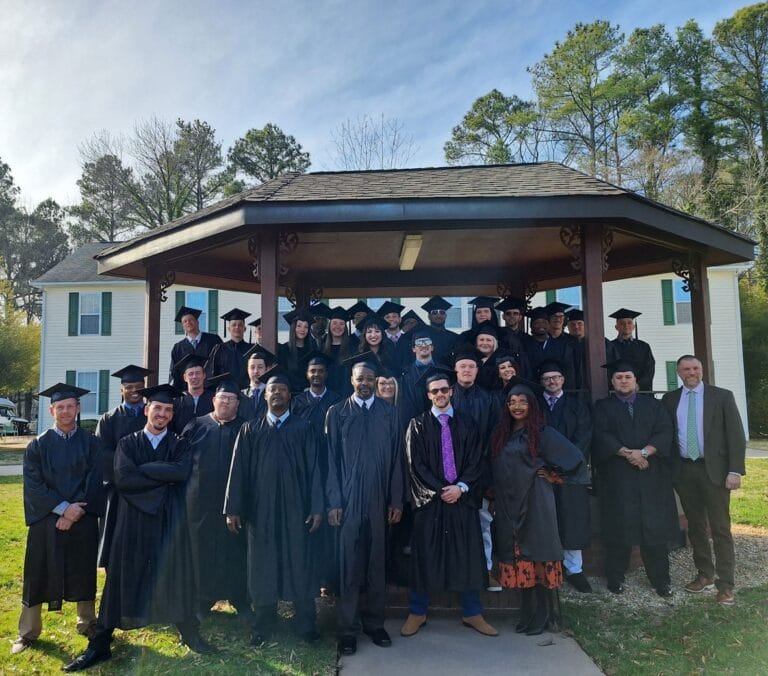 A group of men and women standing and smiling in graduation attire of black cap and gown, gathered together under an outdoor gazebo on the green grass of a campus