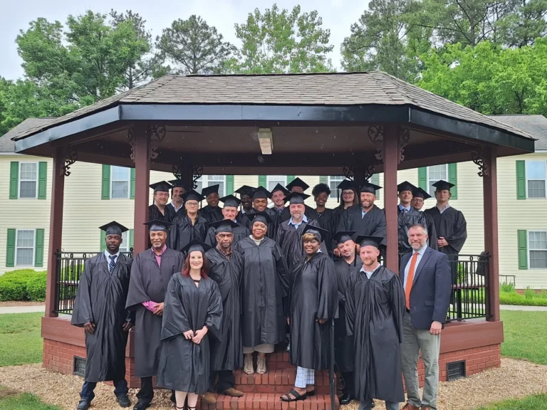 Individuals in graduation cap and gown stand outside under a gazebo on TROSA's campus